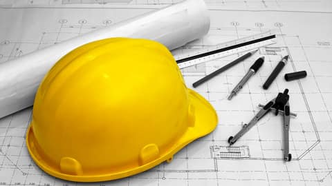 Yellow hard hat on a building plan, with compasses, a ruler and pencils beside it