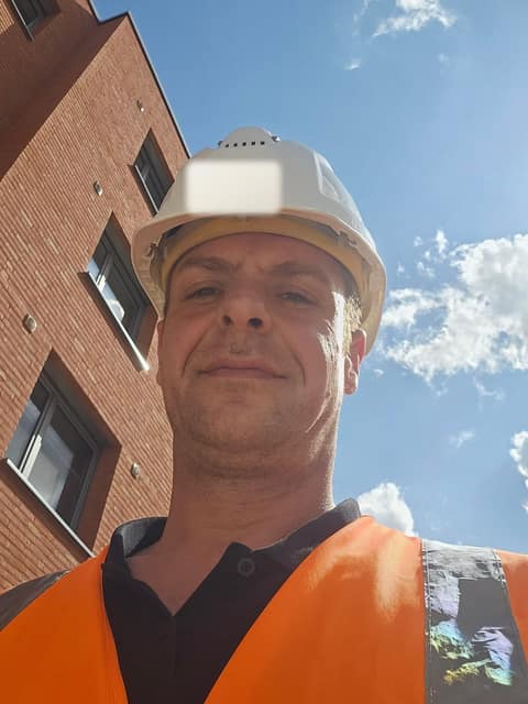 Hannes Timmermann in a high-visibility vest and hard hat on a construction site, a brick facade behind him