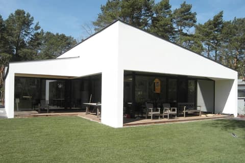 Single-storey white house with a shallow pitched roof and full-height glazing, surrounded by pines