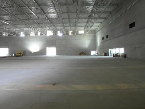 Interior vazio de um grande galpão em obra: paredes de concreto, estrutura metálica do telhado e, ao fundo, uma manipuladora telescópica